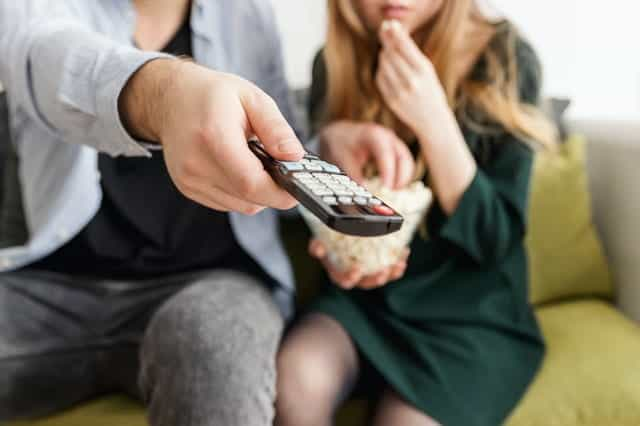 A man and a woman sitting on a sofa, pointing a TV remote and eating popcorn.