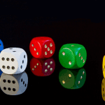 Four die, each one in a different color, laying on a black reflective surface against a black background.