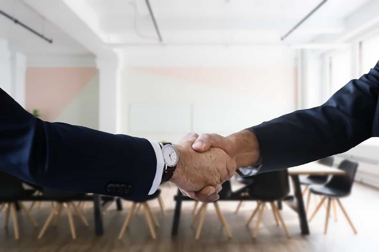 Two hands shaking in front of a long table in a meeting room inside of an office.