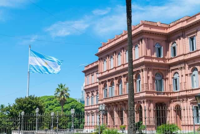 A pink government building, Casa Rosada, in Buenos Aires, with an Argentina flag waving outside on a bright day.