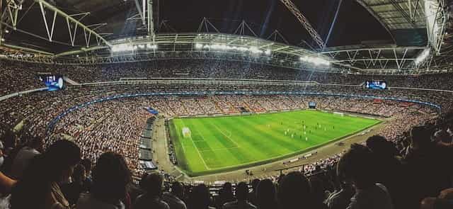 A fully packed-out soccer stadium during a soccer game, with the green pitch in the center, illuminated by bright floodlights.