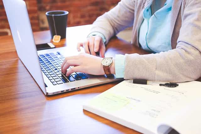 A person sitting in front of a laptop at a desk with a textbook to their left and a smartphone and cup of tea to their right.