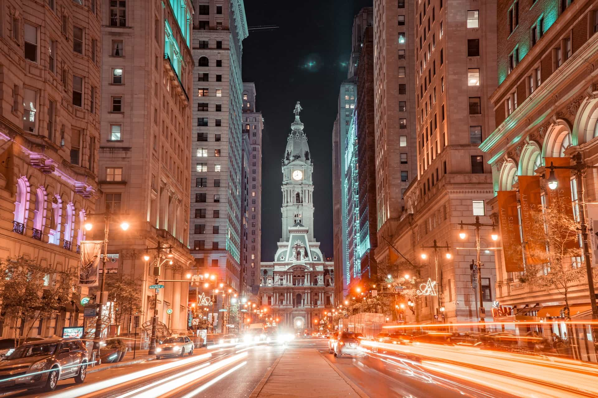 A busy street in downtown Philadelphia, Pennsylvania, featuring a long exposure shot of traffic whizzing by and a tall clock tower at the end of the street.