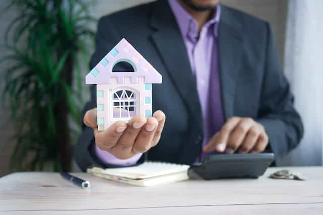 A man in a suit holding a small model house in the palm of his hand.